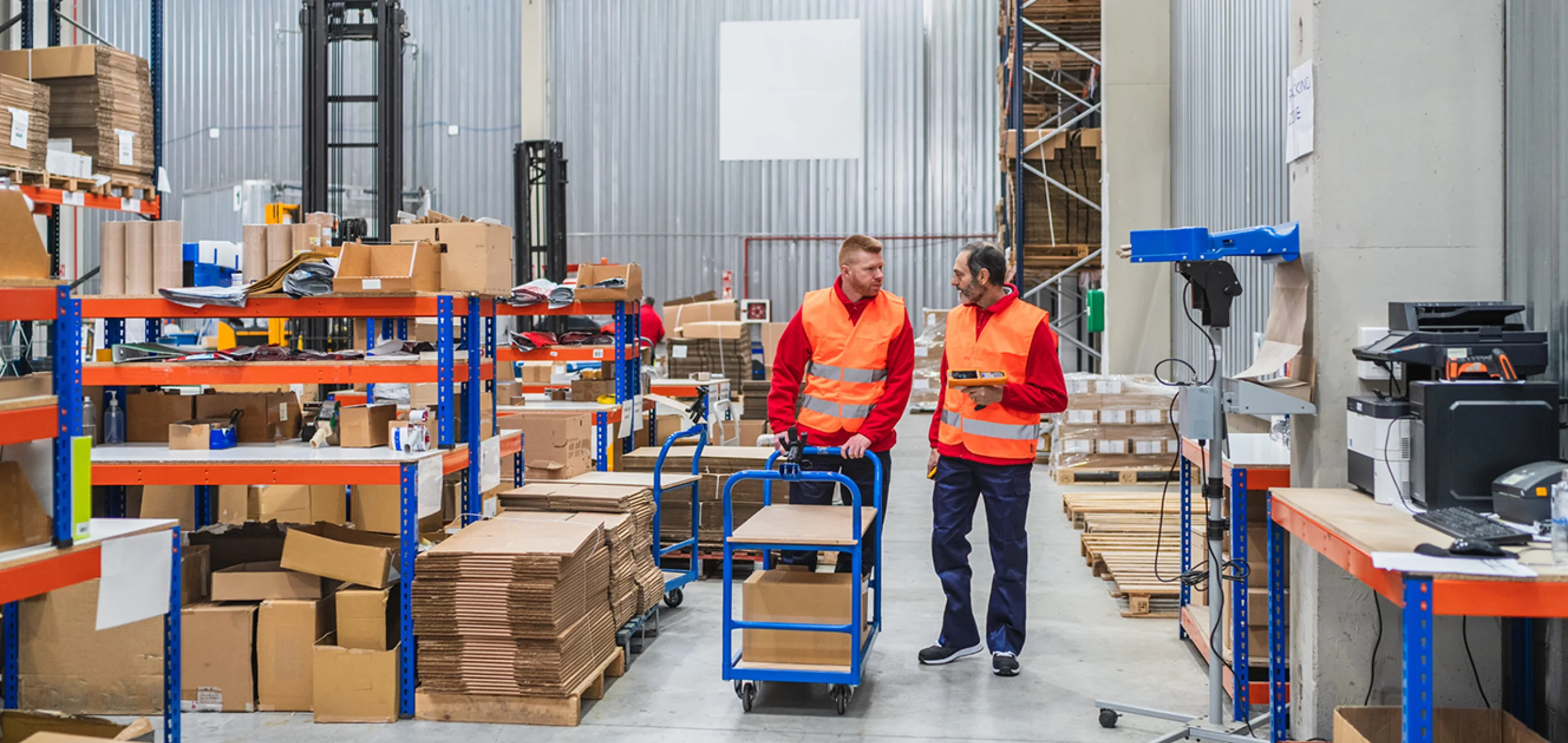 Two men in conversation while pushing a trolley through a warehouse.